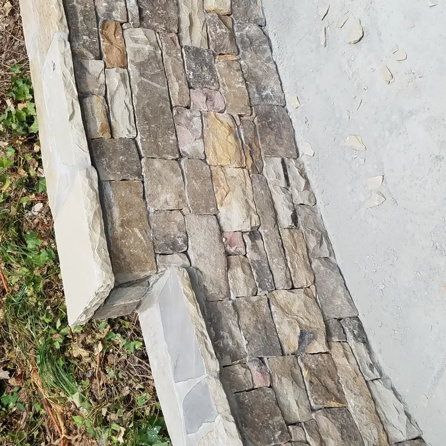 Close-up of stacked stone wall with mixed beige and gray stones next to vegetation and concrete surface.