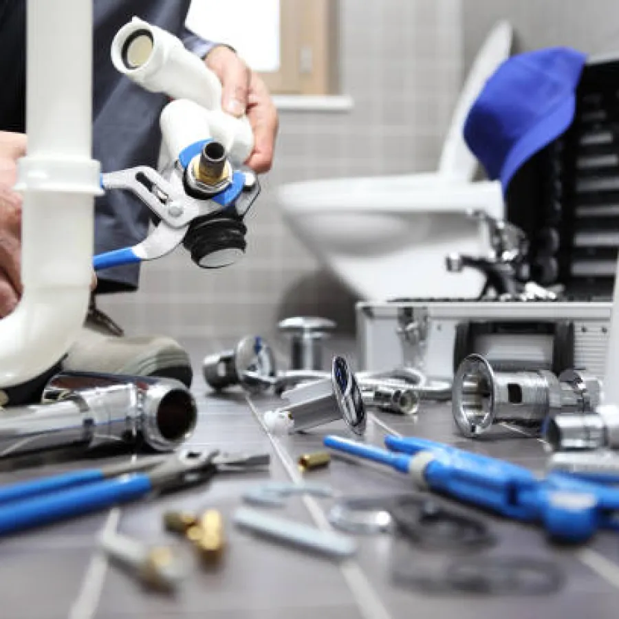 Plumber installing a white PVC pipe with various plumbing tools and fittings on a bathroom floor near a toilet.