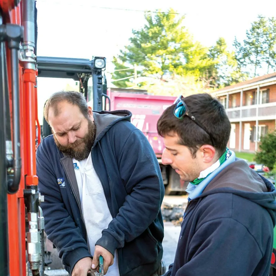 Two men in hoodies working together to fix a metal pipe with a tool beside a red vehicle outdoors.