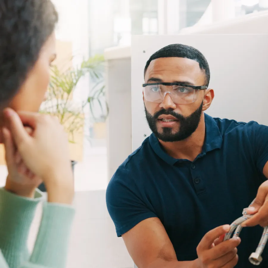 Plumber wearing safety glasses explaining a repair to a woman while fixing pipes under a sink.