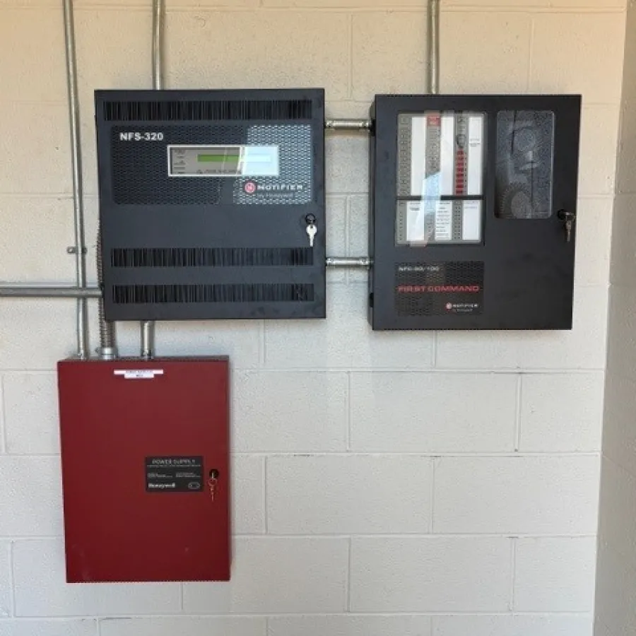 Wall-mounted fire alarm control panels and power supply boxes with metal conduit wiring on a beige brick wall.