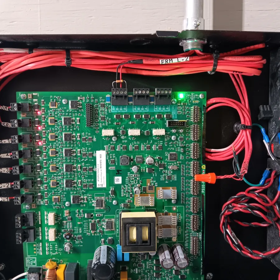 Green fire alarm control panel circuit board with multiple red wires and Power Sonic battery inside black enclosure.