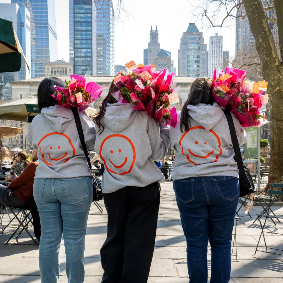 Three people in gray hoodies with orange smiley faces holding pink flower bouquets in a city park with skyscrapers.