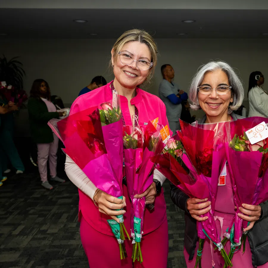 Two smiling women in medical uniforms holding multiple bouquets of pink flowers indoors with people in the background