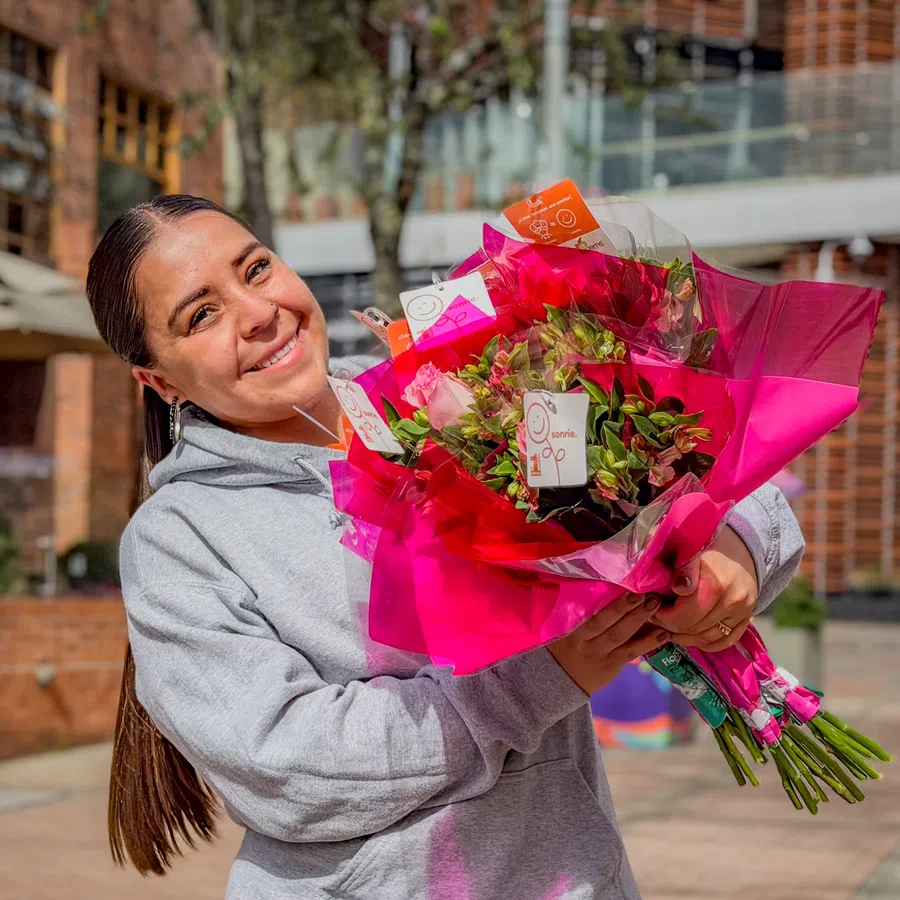 Smiling woman in gray hoodie holding vibrant flower bouquets wrapped in pink and clear plastic outdoors.