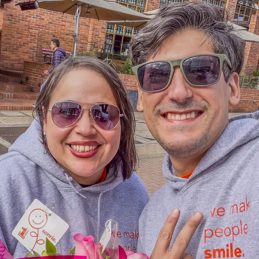Smiling couple in gray hoodies holding pink flower bouquets outdoors under sunny sky