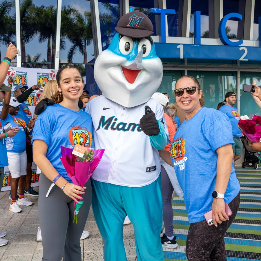 Two women posing with the Miami Marlins mascot outside a stadium, holding flowers at a community event.