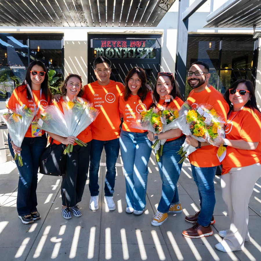 Group of seven people in orange shirts holding flower bouquets smiling outdoors under striped shade.