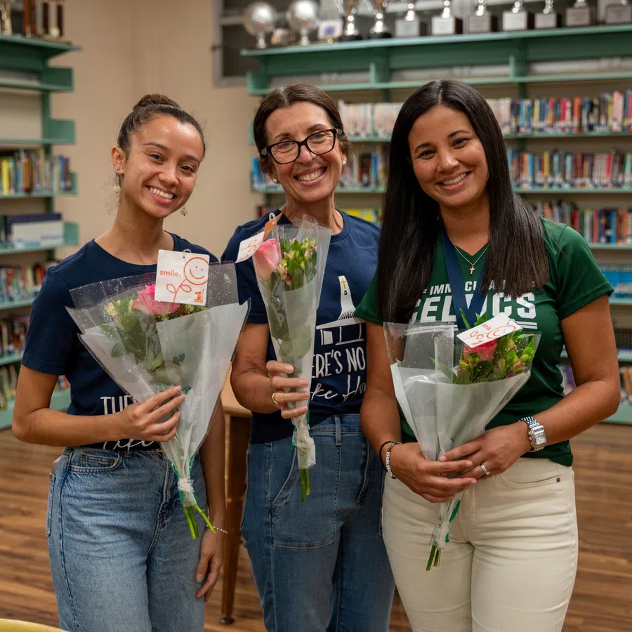 Three women smiling in a library holding wrapped flower bouquets and gift cards.