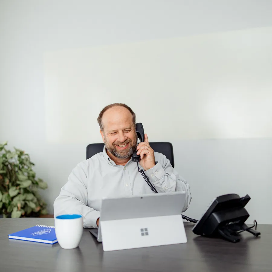 Smiling businessman talking on office phone while working on laptop at desk with notebook and coffee cup