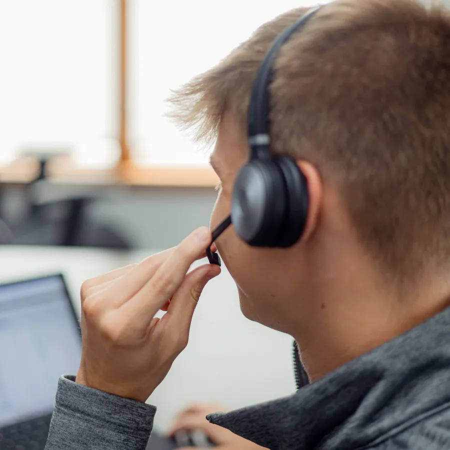 Young man wearing headset with microphone working on laptop in bright office environment.