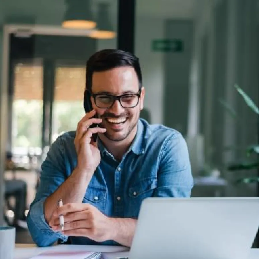 Smiling man in glasses talking on phone at desk with laptop and notebook in modern office.
