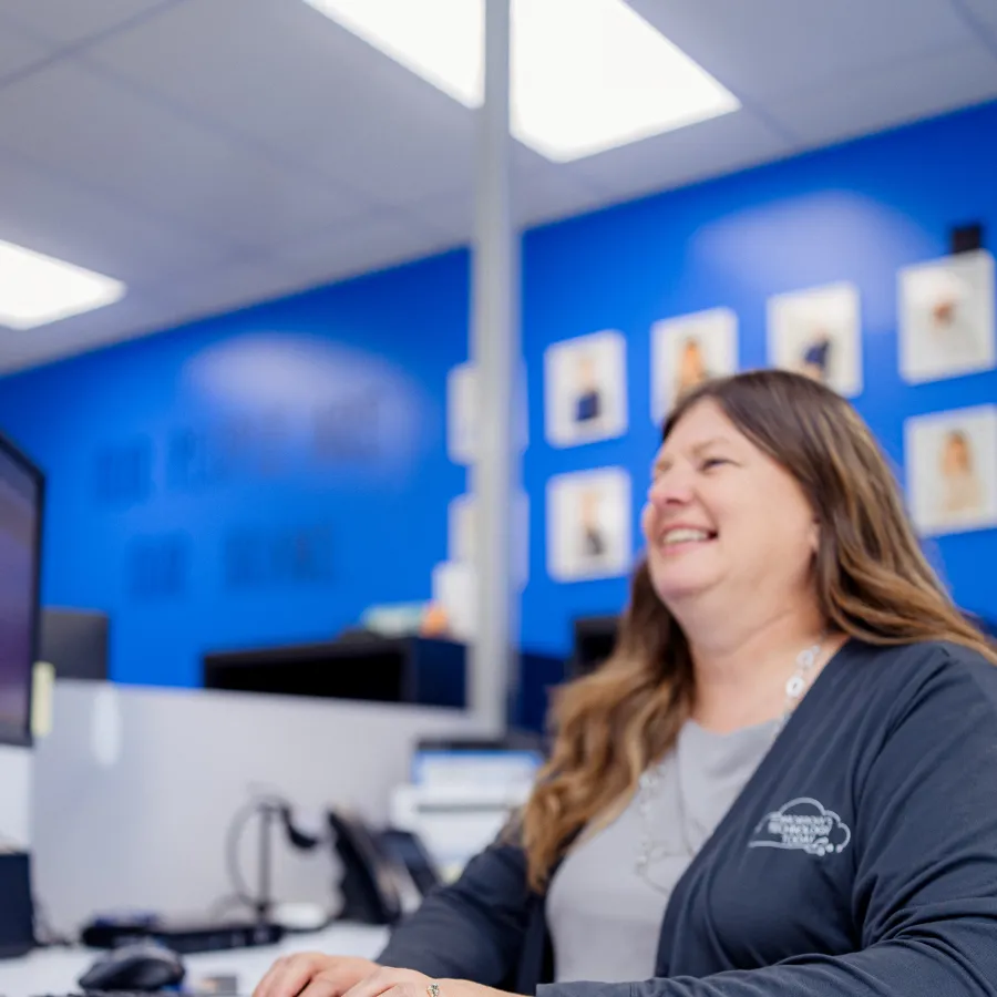 Woman smiling while typing on a keyboard at a white desk in a blue office with framed photos on the wall.