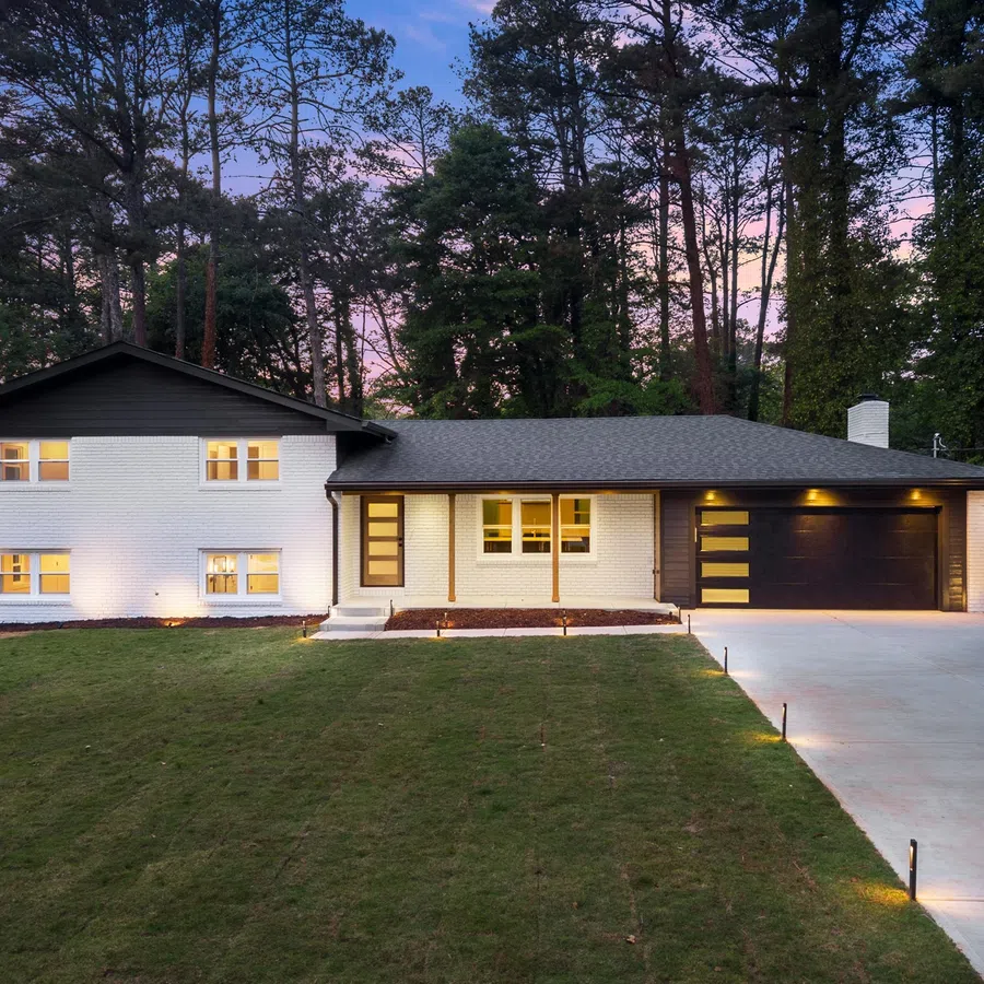 Modern two-story white brick house with black roof and garage at dusk surrounded by tall pine trees and lit driveway.