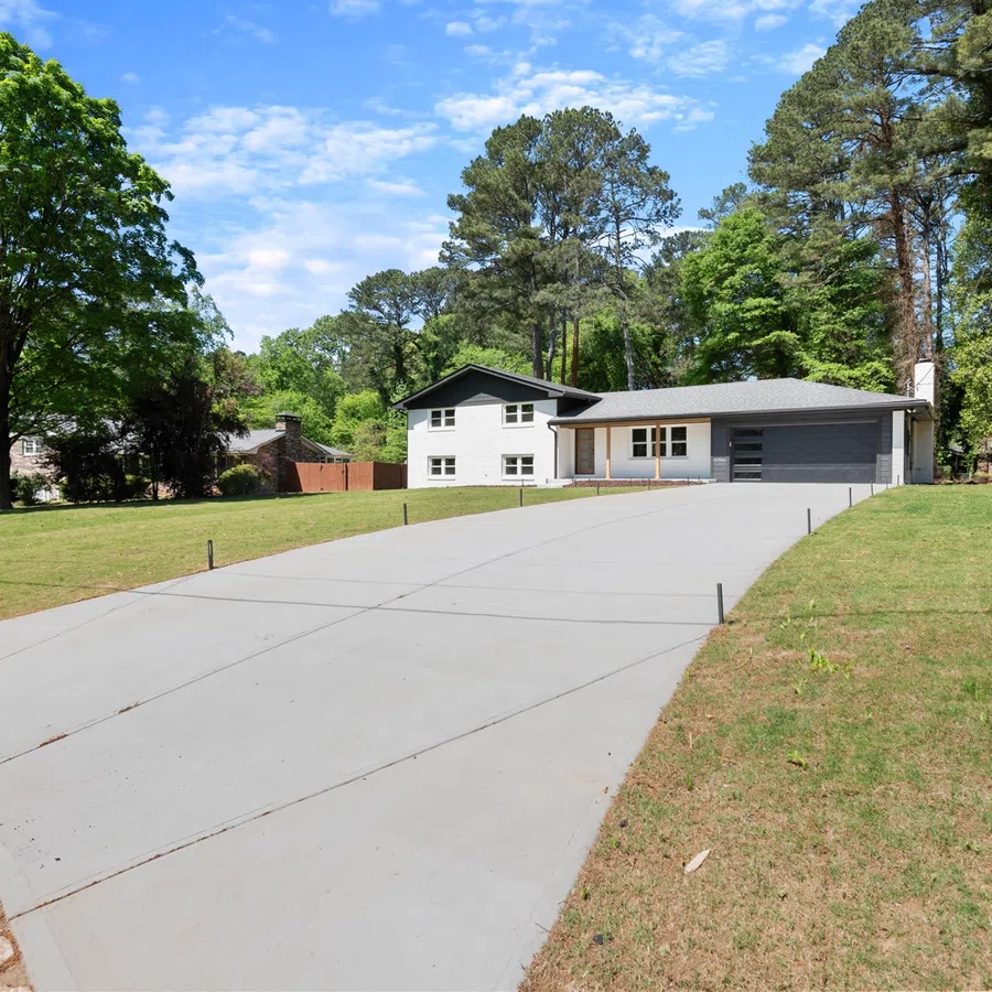 Modern single-story house with a long concrete driveway surrounded by green lawns and trees on a sunny day.