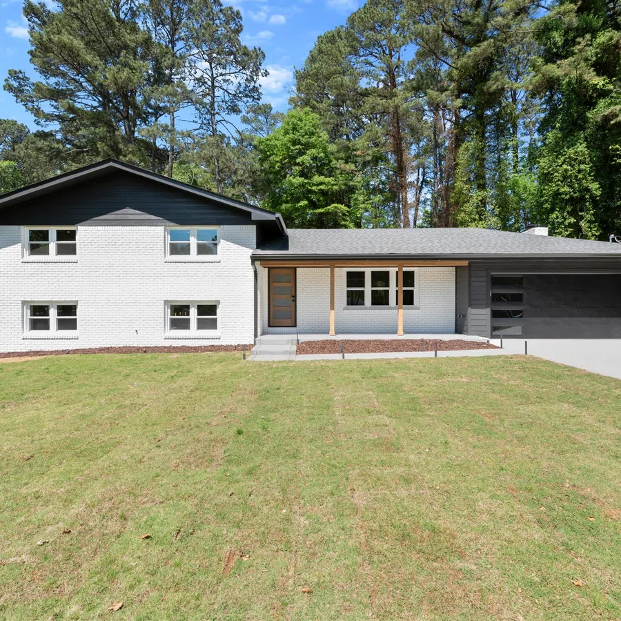 Modern split-level house with white brick exterior, black roof, large green lawn, and surrounding tall trees on a sunny day