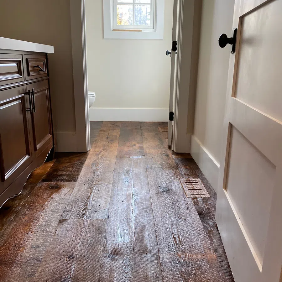 Rustic wooden floor leading to a bright bathroom with white walls and a window letting in natural light.