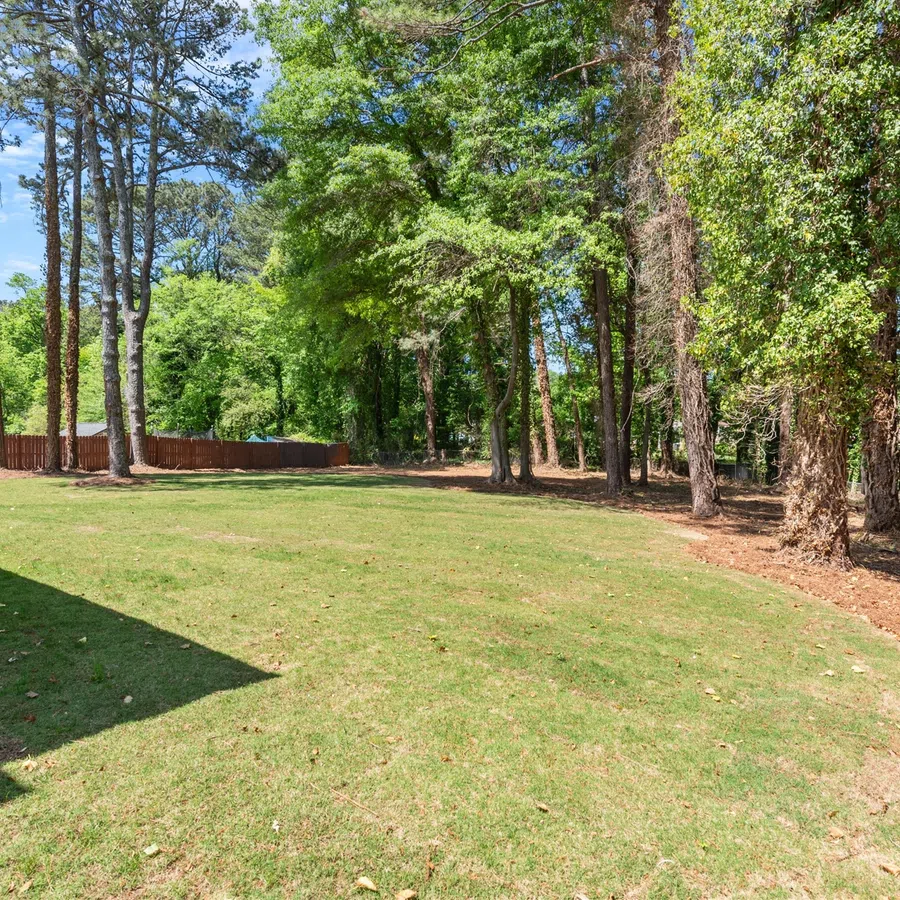 Spacious backyard with green lawn, trees along fence, and shaded porch area on a sunny day.