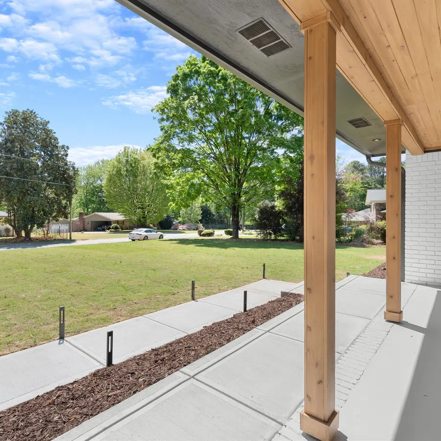 Spacious modern front porch with wooden posts, concrete path, and green lawn under a blue sky with scattered clouds.