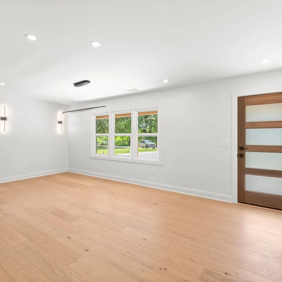 Bright modern empty room with light wood flooring, white walls, large window, dark cabinets, and wooden door.