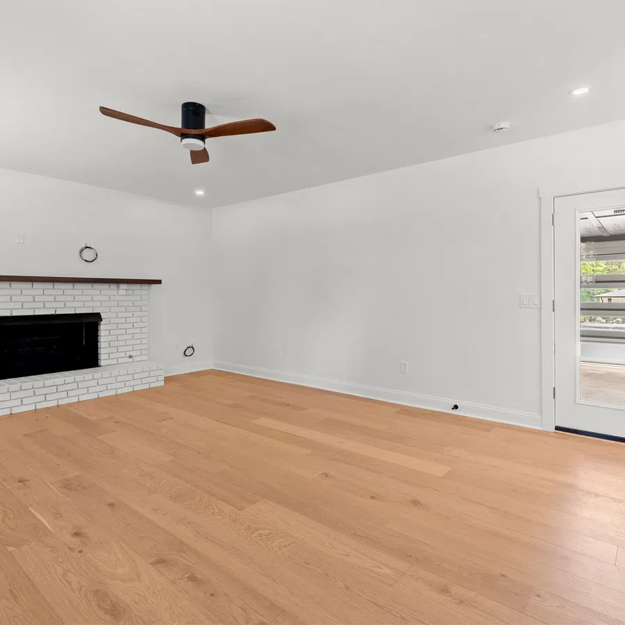 Empty modern living room with light hardwood floors, white walls, brick fireplace, ceiling fan, and glass door.
