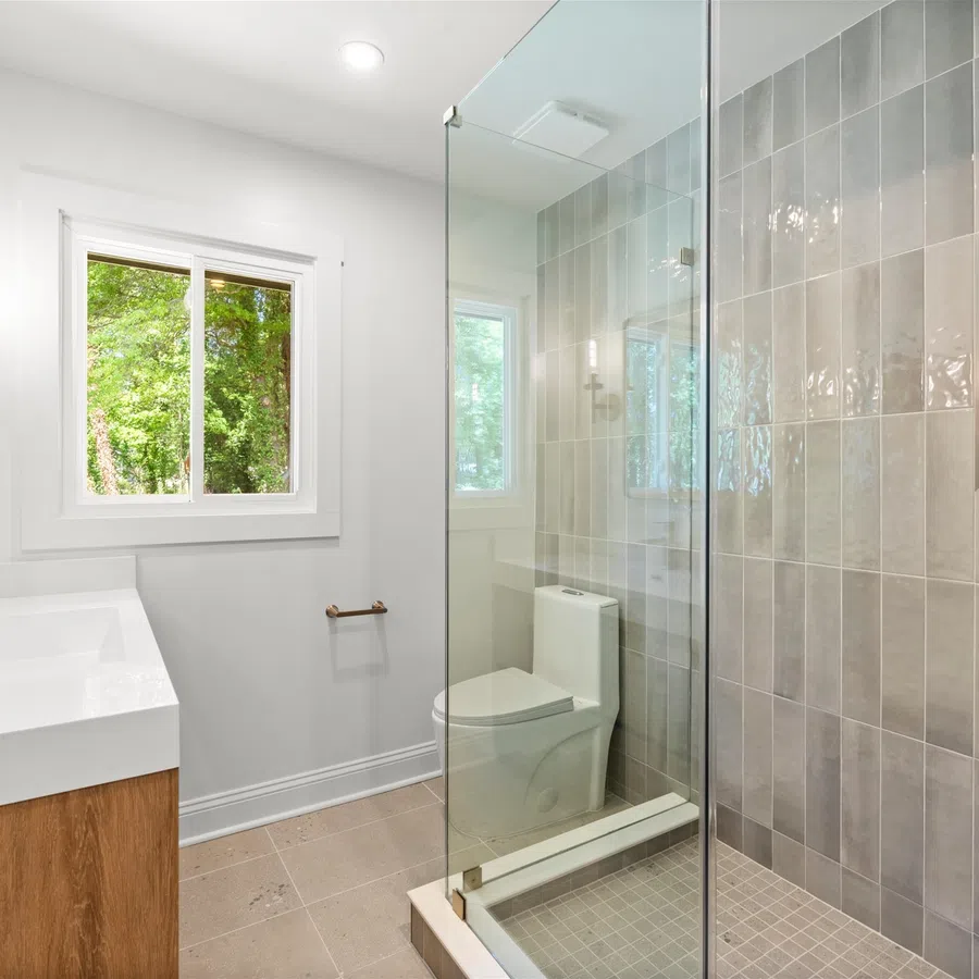 Modern bathroom with glass shower enclosure, wood vanity, wall sconce lighting, and window showing green trees.
