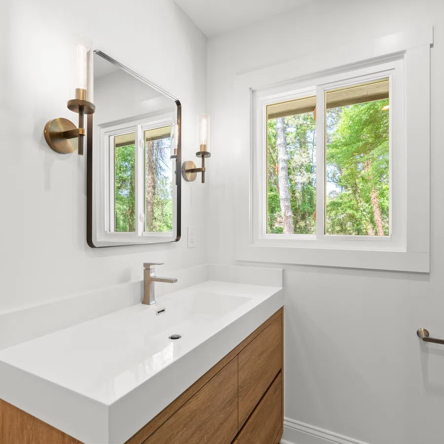 Modern bathroom with white countertop, wooden vanity, rectangular mirror, wall sconces, and window overlooking greenery.