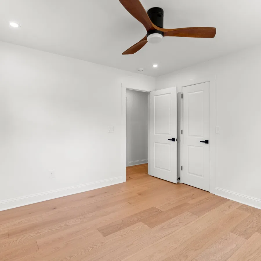 Empty modern bedroom with white walls, light wood flooring, ceiling fan, and two white doors open.