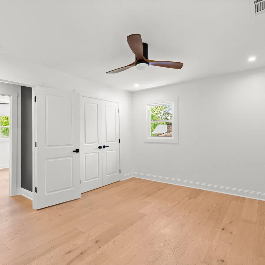 Empty modern bedroom with light wood floors, white walls, ceiling fan, and double closet doors.