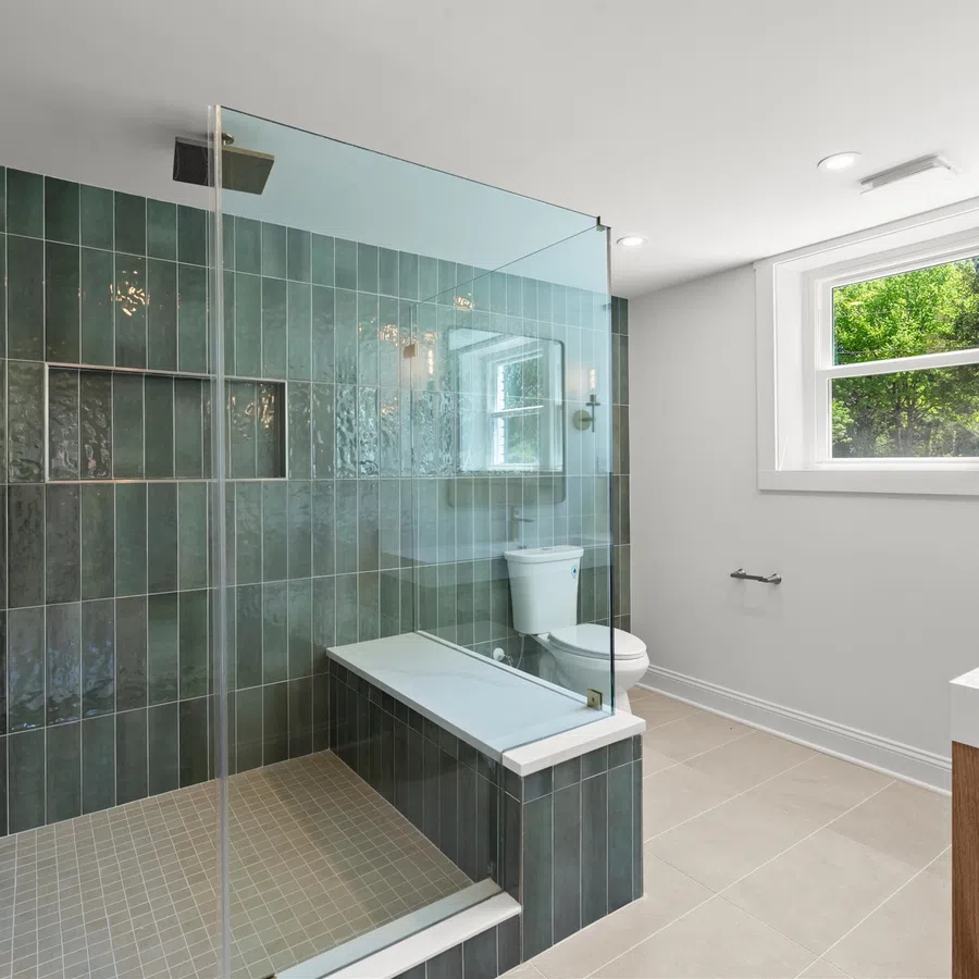 Modern bathroom with green tiled walk-in shower, built-in bench, white toilet, and large window with natural light.