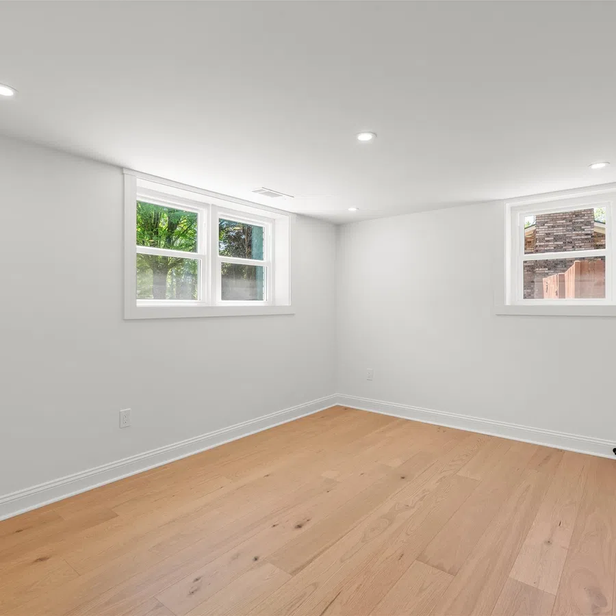Bright empty room with white walls, light wood flooring, two windows, and recessed ceiling lights.