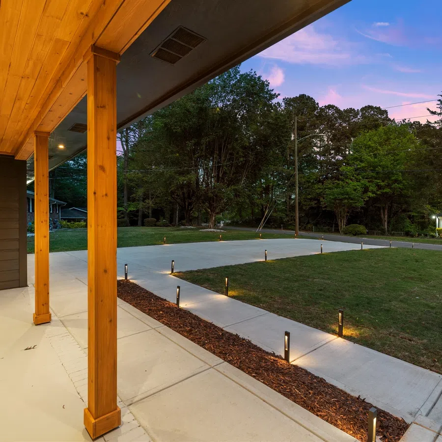 Covered wooden porch with recessed lighting overlooking a lit concrete driveway and green yard at dusk.