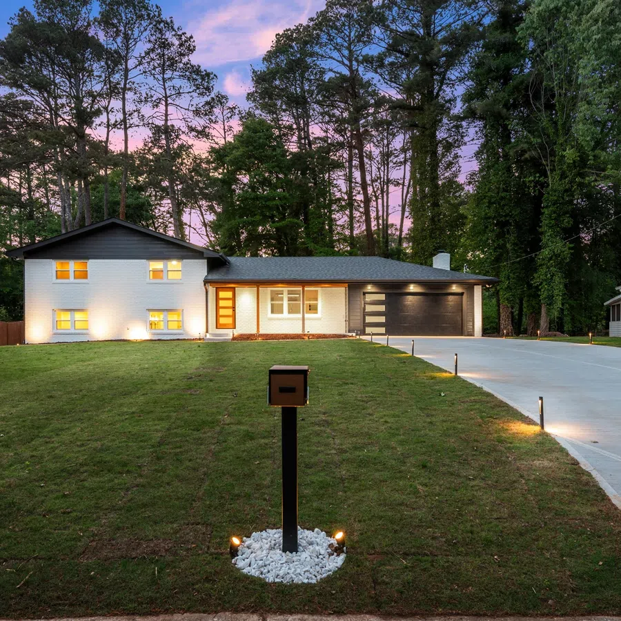 Modern split-level home with illuminated windows, wide driveway, and mailbox on a spacious green lawn at sunset.