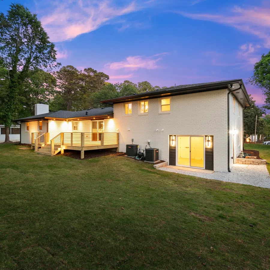 Modern two-story white brick house with wooden deck, large backyard, and glowing interior lights at sunset.