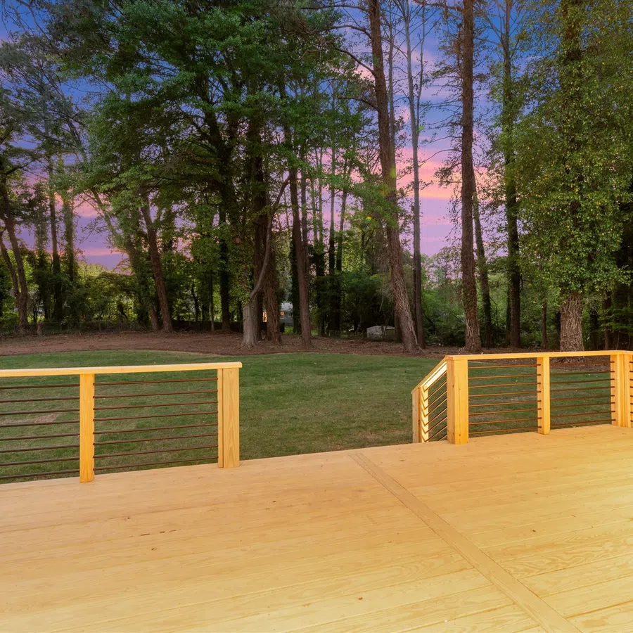 Spacious wooden deck with metal railing overlooking a green backyard and tall trees at sunset.