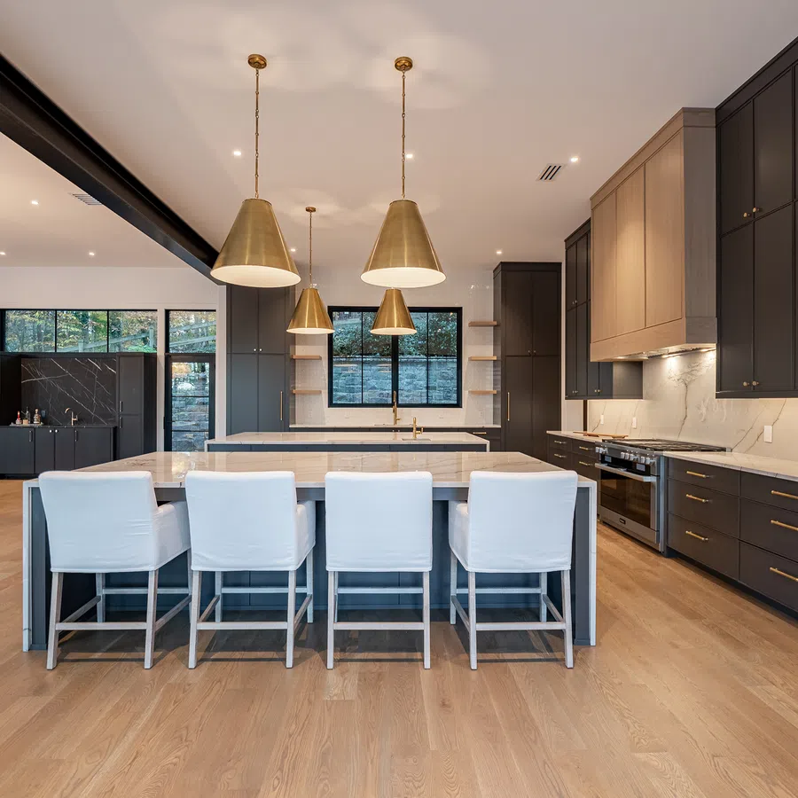Modern kitchen with marble island, dark cabinetry, brass pendant lights, and white cushioned bar stools on wood floor