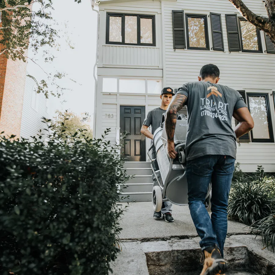Two workers carrying a large appliance up the steps to a white two-story house on a sunny day.