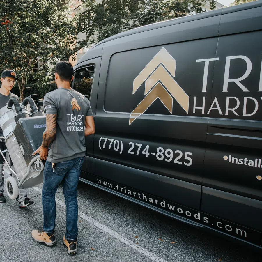 Two workers unloading equipment from a Tri Art Hardwood van in an outdoor setting with trees and pavement.