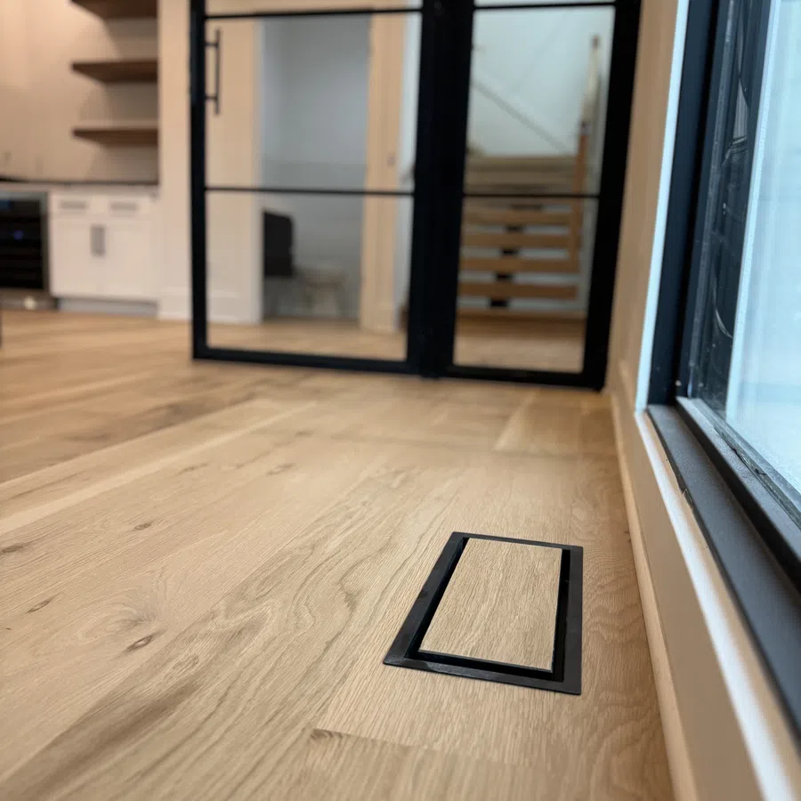 Modern wooden floor with a hidden floor vent near a glass door leading to stairs and kitchen area.