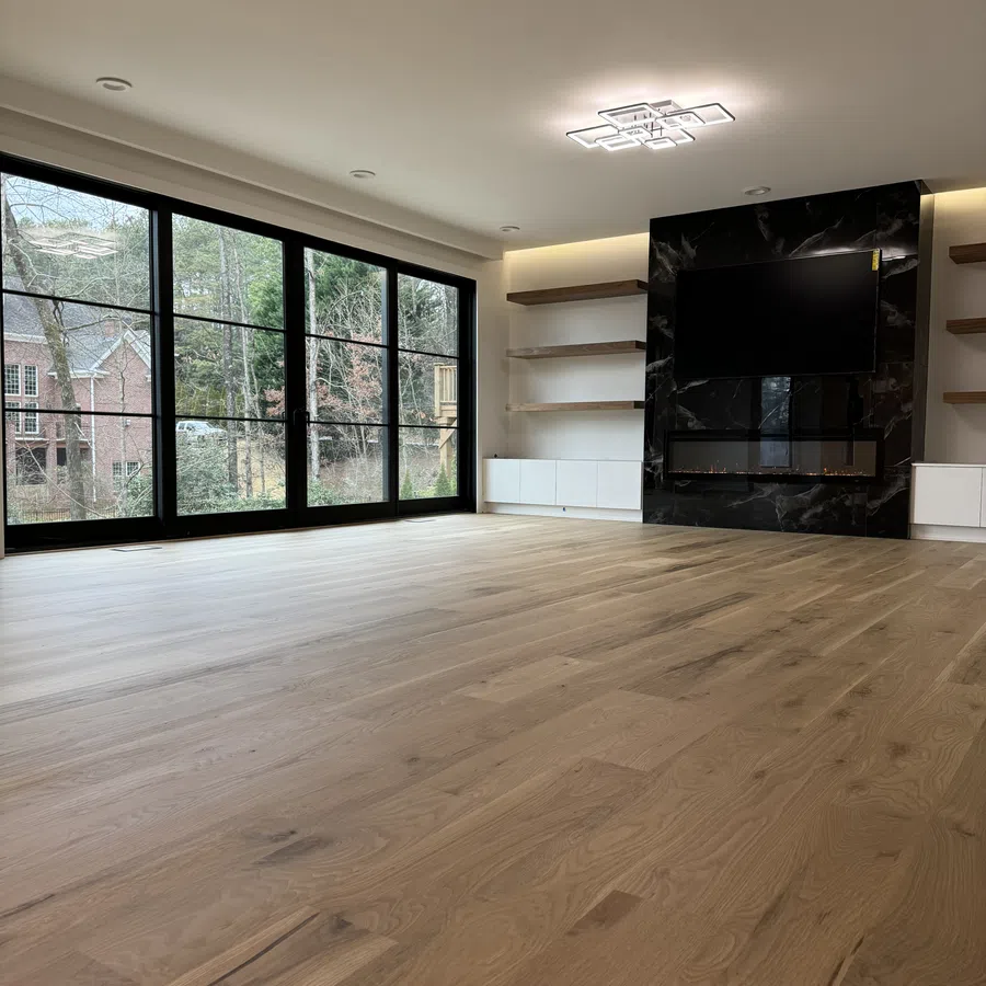 Modern empty living room with light wood flooring, large black-framed windows, wall-mounted TV, and fireplace.