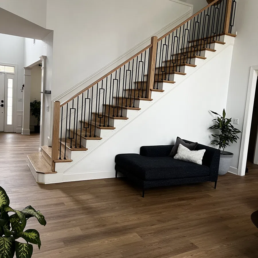 Modern living space with wooden staircase, black chaise lounge, white walls, and wood flooring with plants.