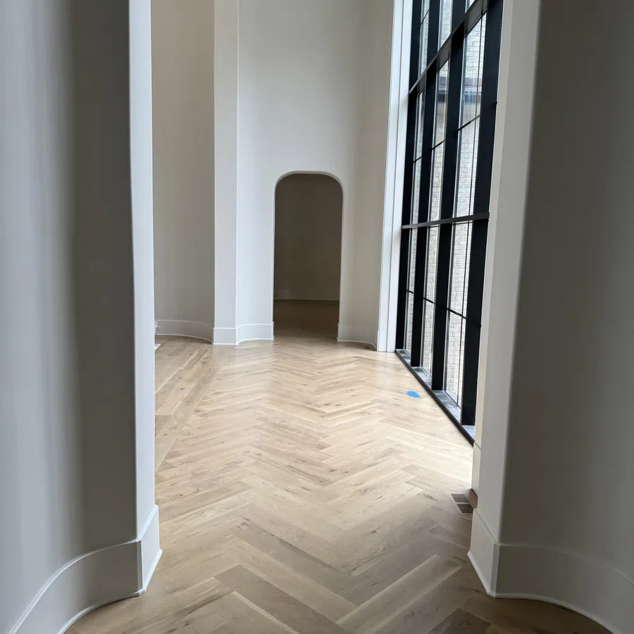 Bright hallway with light wood herringbone floor, white walls, and tall black-framed windows letting in natural light