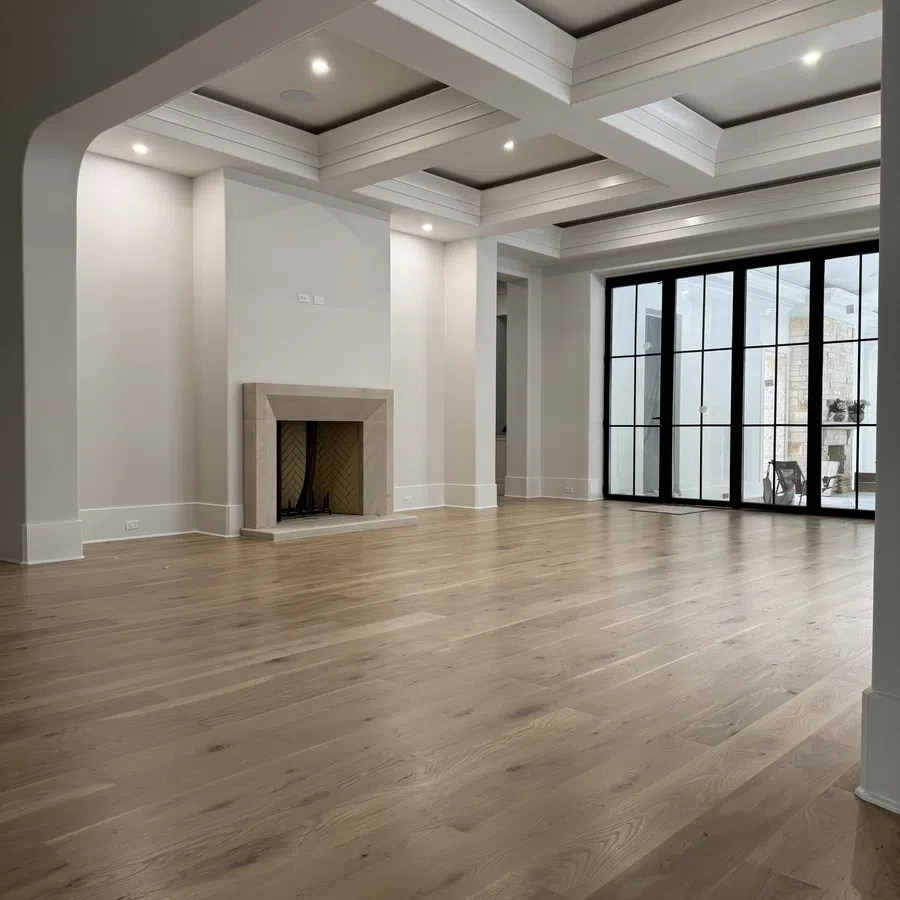 Spacious empty living room with hardwood floors, white walls, coffered ceiling, fireplace, and large glass doors.
