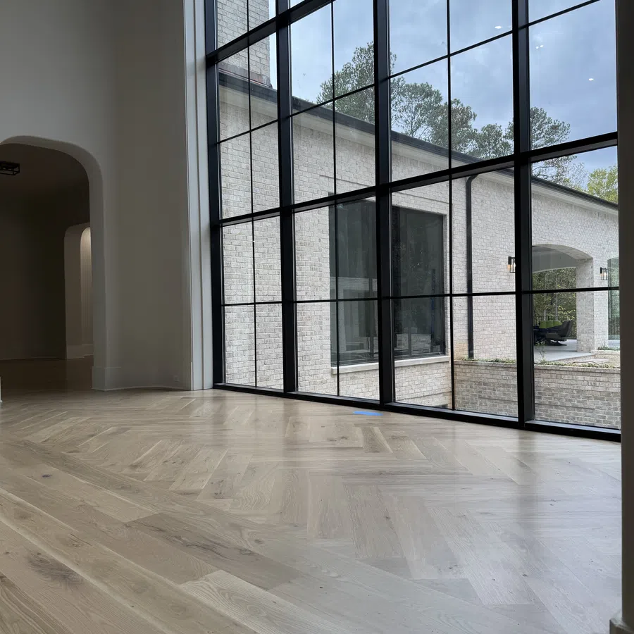 Modern empty room with large black-framed floor-to-ceiling windows and light wood parquet flooring.