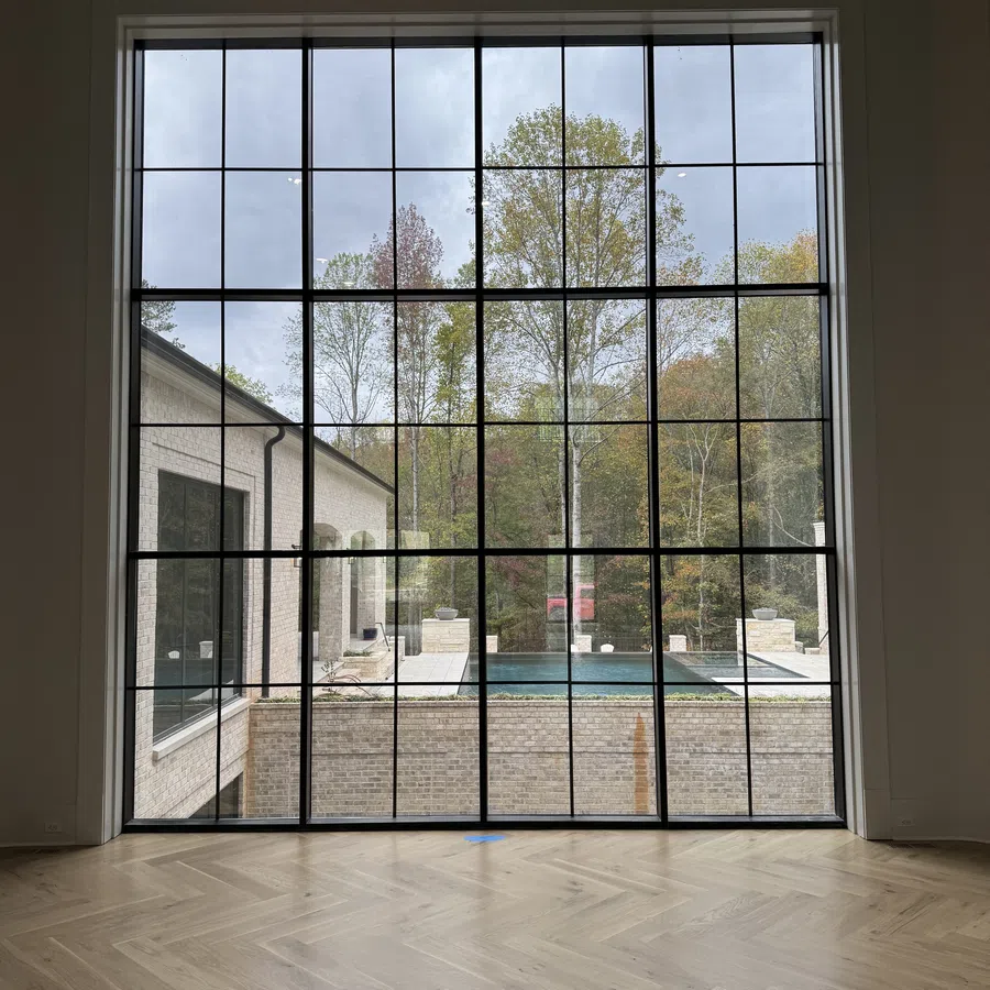 Large floor-to-ceiling window with black grid framing overlooking a backyard pool and trees outside under cloudy sky.