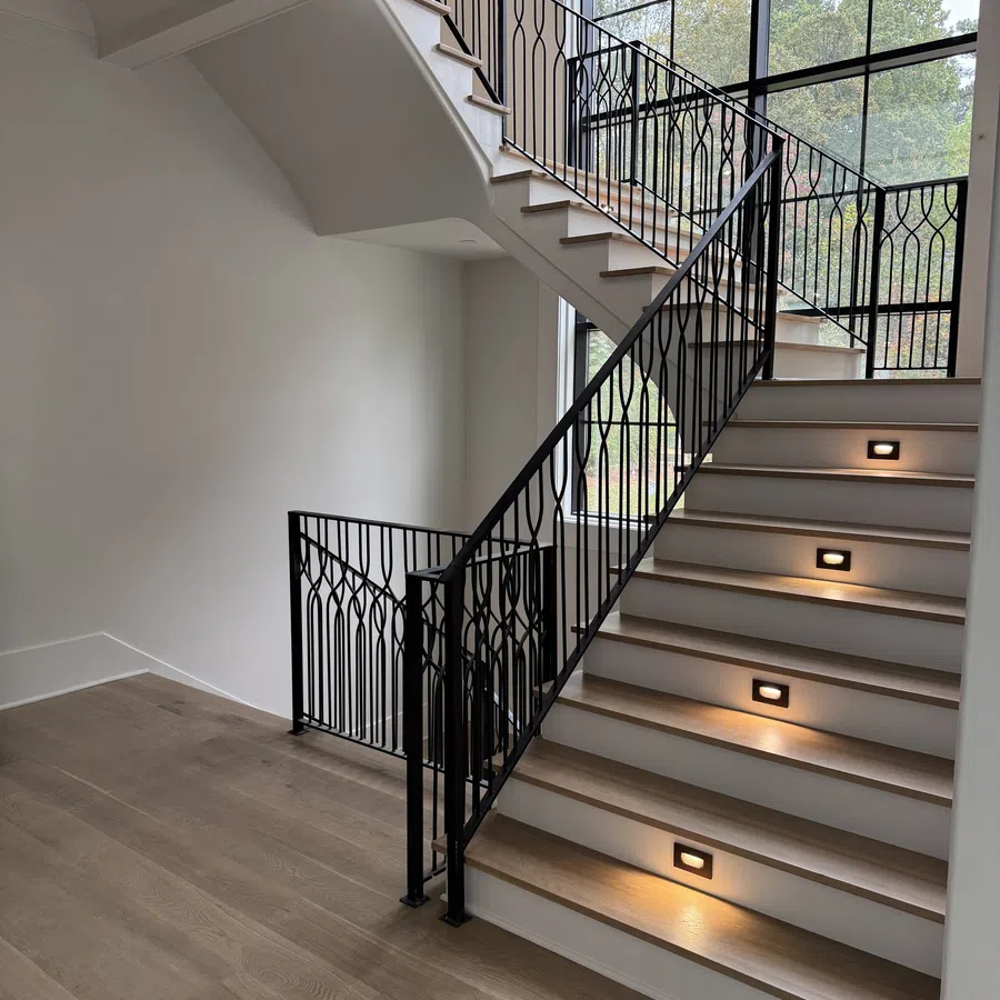 Modern staircase with black metal railing, wooden steps, and built-in step lighting by large windows.