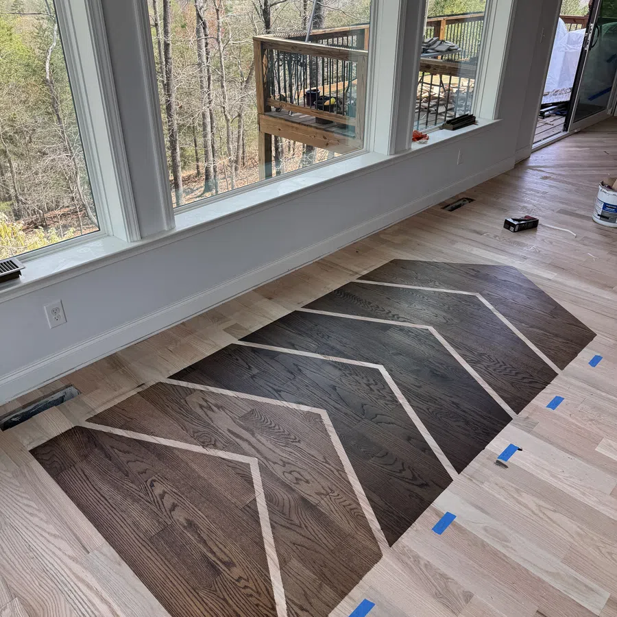 Partially installed geometric dark wood floor pattern in light wood room with large windows and outdoor deck view.