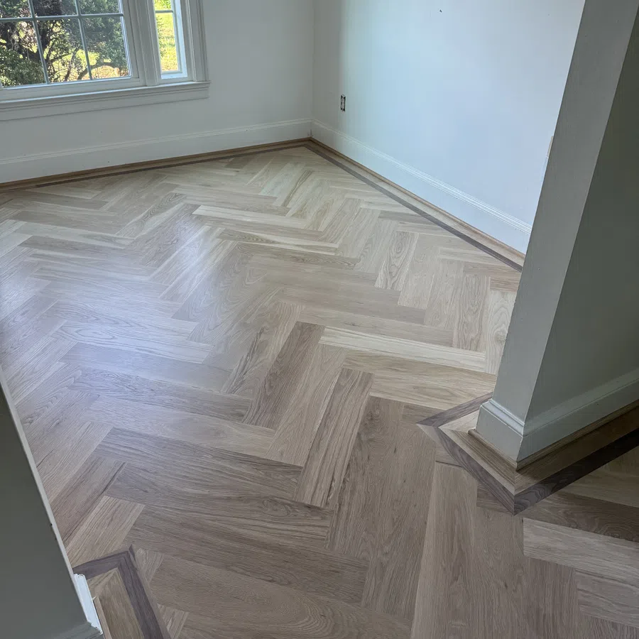 Light wood herringbone pattern flooring with dark wood border in an empty room with white walls and large window.