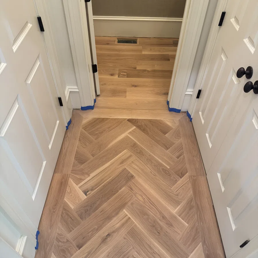 Newly installed herringbone wood floor section surrounded by traditional wood planks in a hallway with white doors.