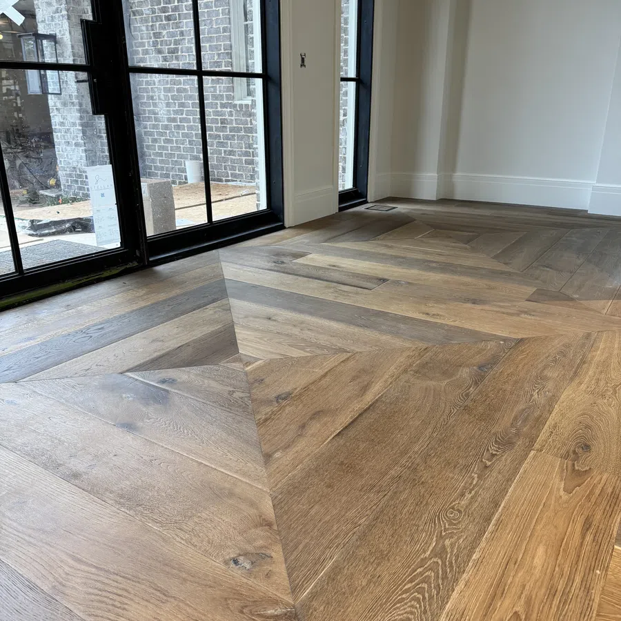 Close-up of herringbone patterned wooden floor near large black-framed windows in a modern room.